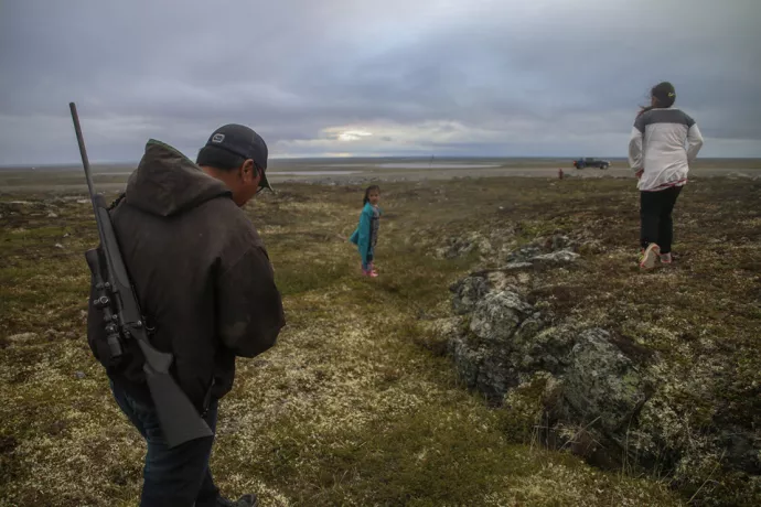 A man wearing a baseball hat with a rifle slung over his back walks along the tundra behind two girls, one stopping to look back, with a truck pickup truck in the background