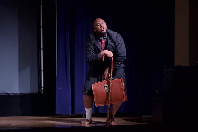 Actor standing on stage in a blue suit, pant legs rulled up, wearing a bow tie and leaning on a cane while holding a battered briefcase