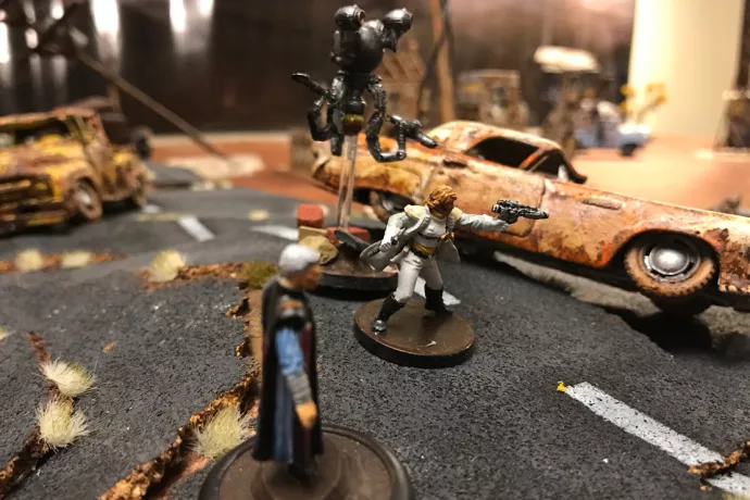 Photo of three miniature humanoid figures on an apocalyptic tabletop scene scattered with debris and overturned cars.