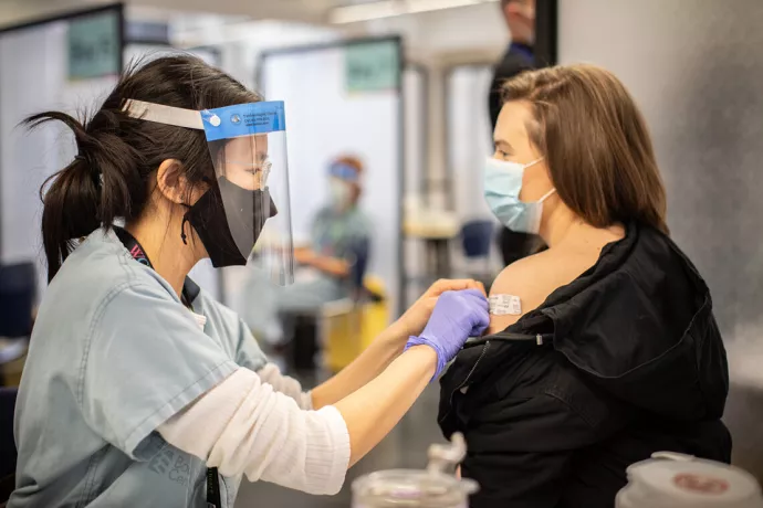 Woman wearing face mask, face shield, blue scrubs and purple latex gloves puts a band-aid on the upper arm of another woman at a vaccine clinic