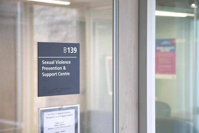 A glass door with a sign on it that reads: B139 Sexual Violence Prevention & Support Centre. Sign below reads: Tri campus sexual violence prevention and support. Office hours: 9am-5pm Monday, Tuesday, Thursday, Friday (rest of sign cut off from view)