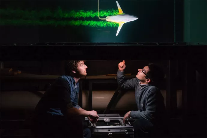 Two men sitting under a table, one pointing up. Top of table shows a small rocket-like device blasting buy with green smoke trailing behind