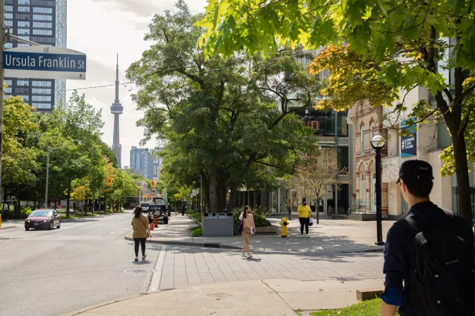 People wearing face masks walking along tree-lined street with the CN Tower in the background. Street sign reads Ursula Franklin St.