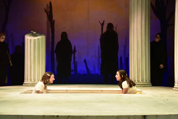Two girls sit in a rectangular pit on stage, facing one another as if in discussion. In the background are shadows of people and a broken picket fence