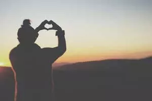 Woman holds hands in shape of a heart with sunset in the background