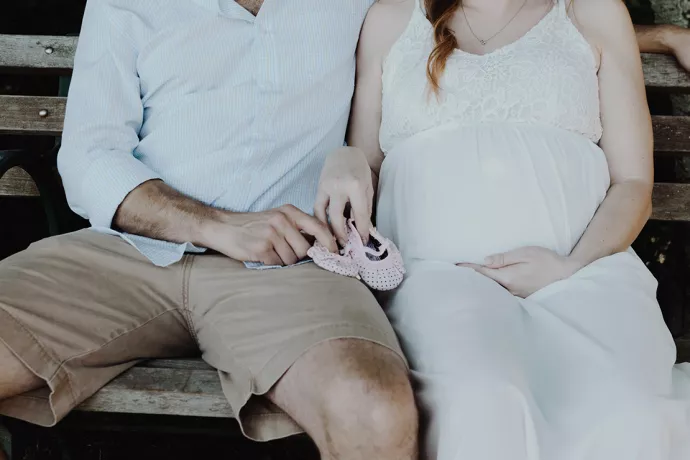 Couple on bench, man wearing white button down shirt and khaki shorts, pregnant woman wearing white dress, both holding a pair of pink baby shoes between them