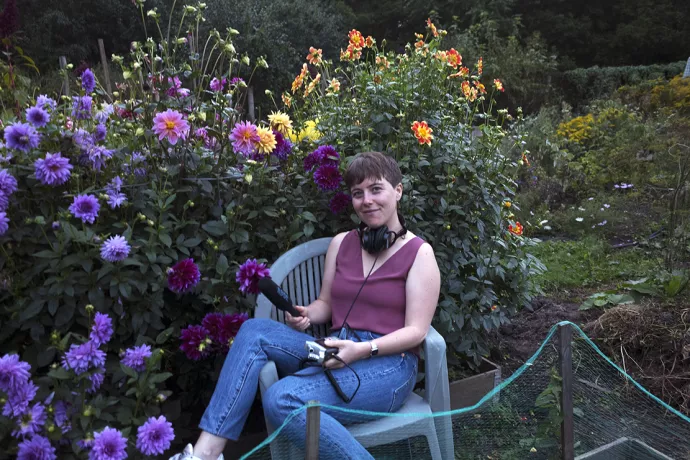 Lauren Ramsay sitting in a patio chair in a garden surrounded by bushes and flowers, wearing jeans and a sleeveless purple shirt, holding a microphone, audio recorder and wearing large headphones around her neck