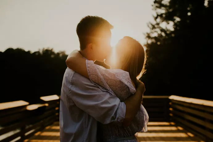 Man and woman standing on a dock kissing, with sunlight streaming behind them, obscuring the view of them kissing