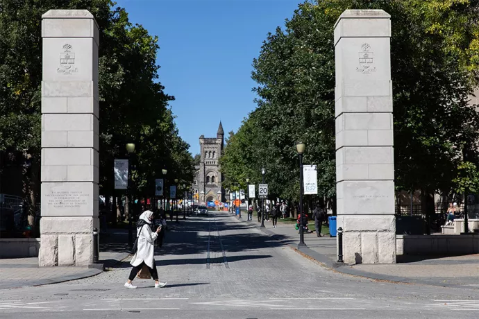 Woman walking past two stone pillars, each bearing a carving of the U of T coat of arms near the top.