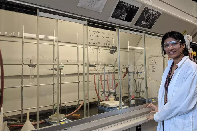Avneet in lab coat standing next to shelves of beakers