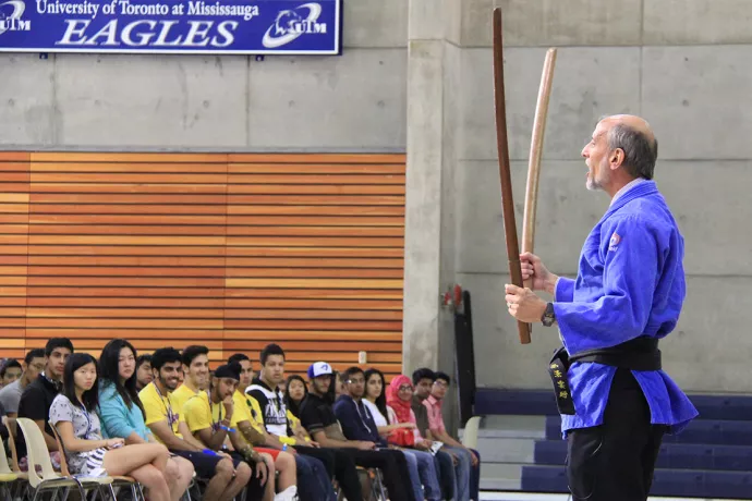 Ulli Krull in karate-style outfit holding swords in front of audience of students in a gym
