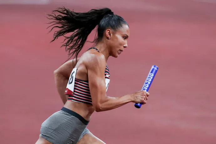 Alicia Brown running, with a paper number on her back and chest, holding a blue baton that reads Tokyo 2020