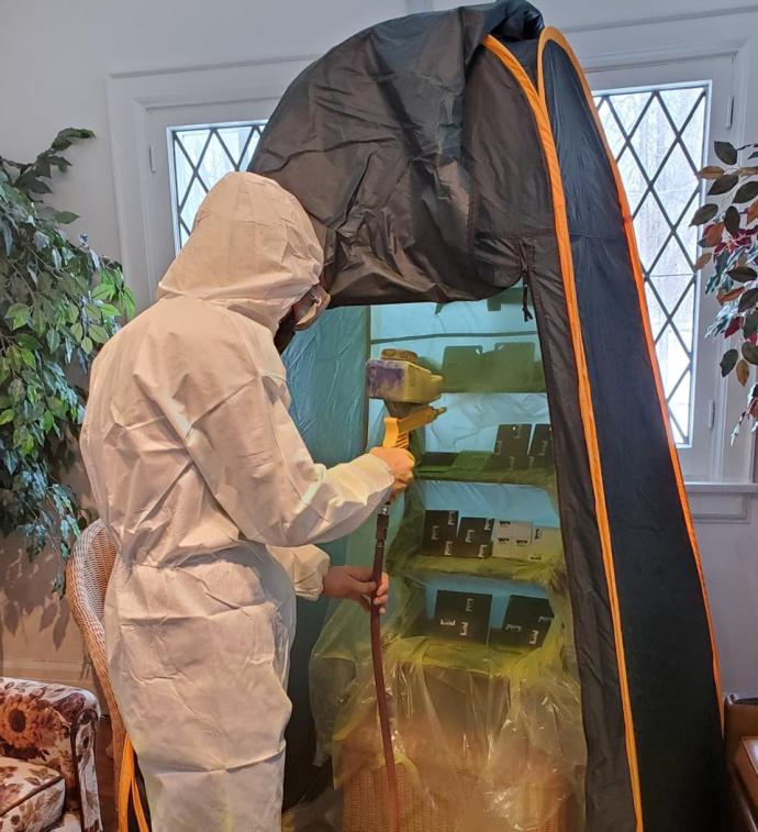 Person in full, white Hazmat-type suit with a handheld sandblaster spraying yellow dust over square samples on shelving unit