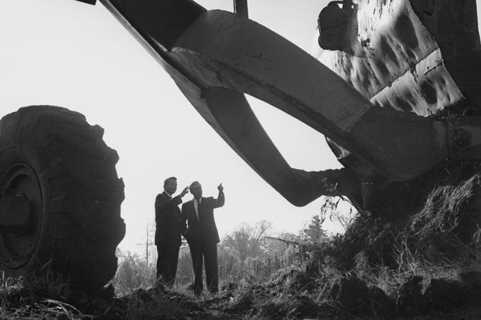 Black and white photo of two men in suits standing in the dirt with a large backhoe-like machine overhead