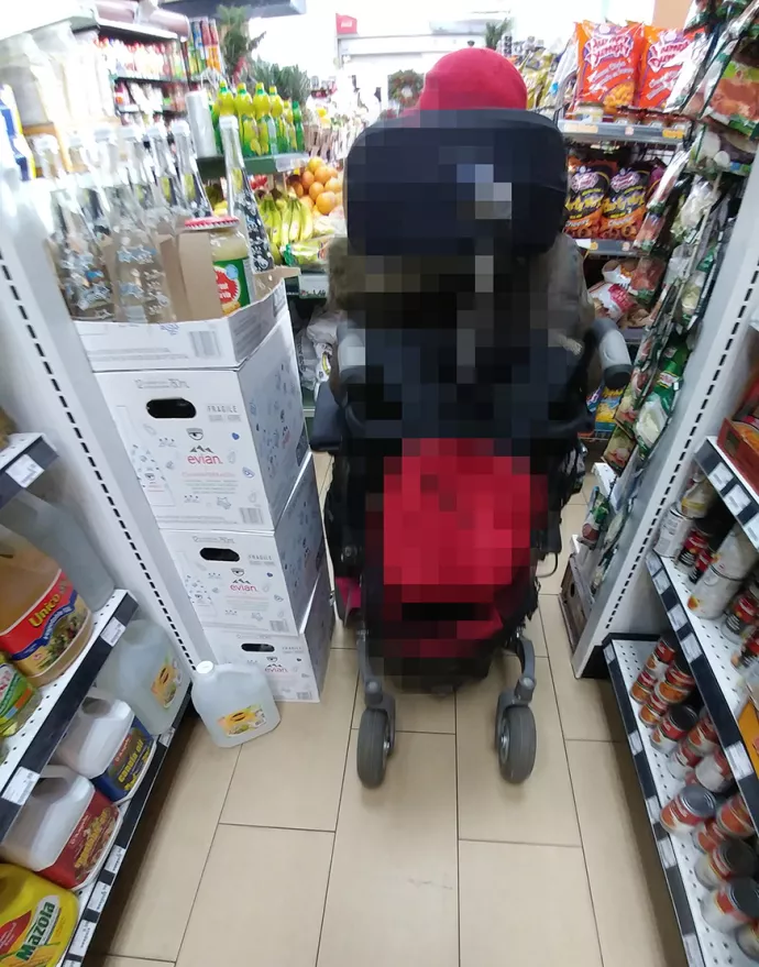 Individual on electic wheelchair seen going down a very narrow grocery aisle, with a display of Evian water blocking part of the aisle. The wheelchair is almost the width of the narrow space provided.