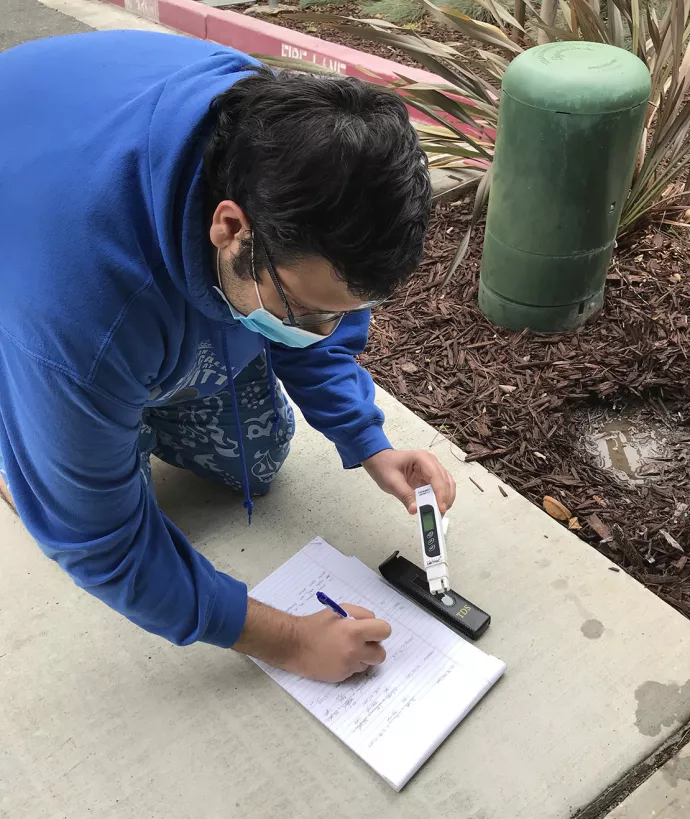 Jamsheed wearing a blue hoodie kneeling on a sidewalk, writing in a notebook with his right hand while his left hand holds a white, rectangular sensor to a black, rectangular device sitting on the ground