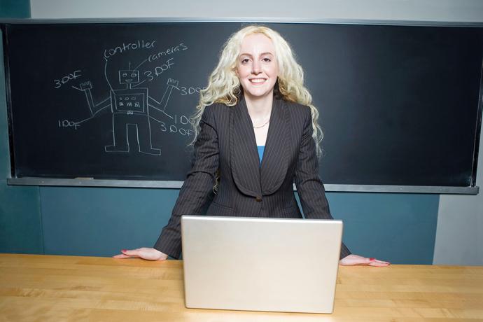 Woman in black pinstripe suit jacket standing at a wooden desk with open laptop, in the background is a chalkboard with a chalk drawing of a robot