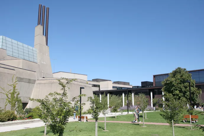 large concrete building with three smoke stacks rising to the left, grass in front with people walking along paths