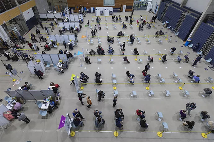 Overhead image of vaccine clinic, rows of chairs in large gym physically distanced with people sitting in them