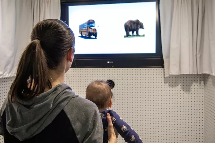 A woman with a baby in her lab sit facing away from the camera, in front of them is a TV with the image of a school bus and a bear on it