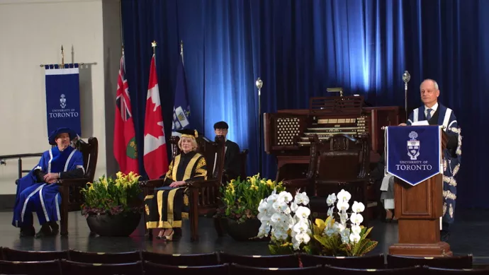 Meric Gertler standing at a podium on the stage at Convocation Hall, wearing a blue robe. Also on stage is Rose Patten, sitting in a large wooden chair on stage.