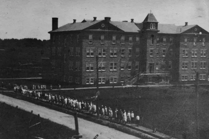 Black and white grainy image of large stone building and line of people leading into the building
