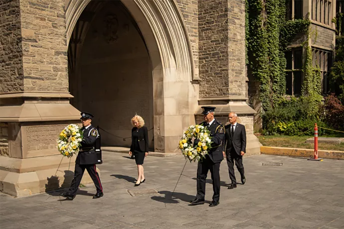 The provost and president walk behind to men carrying large wreaths of yellow and white flowers.