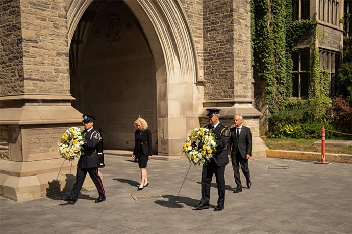 The provost and president walk behind to men carrying large wreaths of yellow and white flowers.