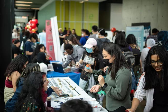 Three students sitting at a table with miniature plastic bones and skulls and bowls of candy talking to students in a crowded corridor during UTM's Club Fair.