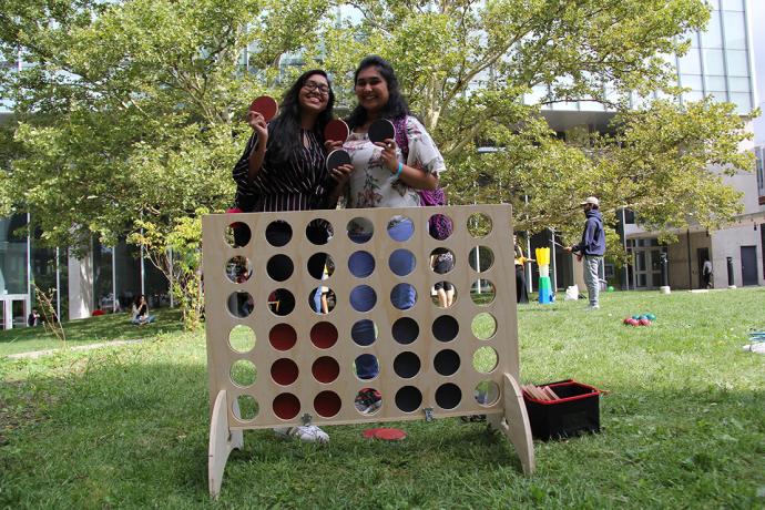 Two students stand in front of a large connect 4 game on a grassy area.