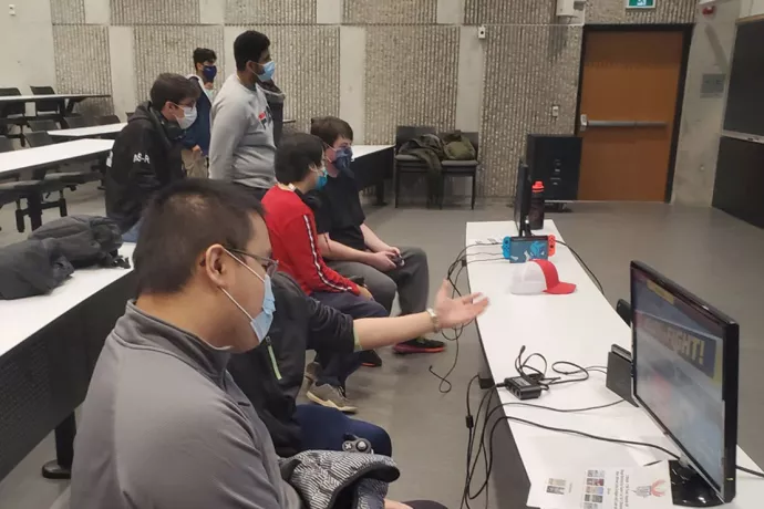 Six students wearing blue face masks sitting in front of a long white table in a lecture hall holding gaming controllers and looking at computer screens.  