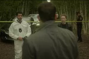 Zach Smadu in a scene from Cardinal. He wears a white hazmat suit and stands with two police officers in a forest setting.