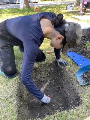 A student kneels over a square hole dug in the ground as she carefully removes dirt from the hole, revealing what looks to be a large tree root.