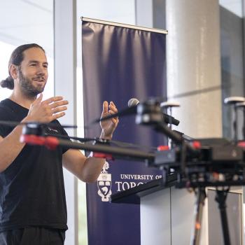 Bryce Jones standing at a podium speaking, next to him is a blue sign that reads University of Toronto Mississauga and in the foreground is a drone
