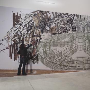 Large painting on wall in gallery showing a woman in jeans and a black shirt with a paint brush painting black, abstract lines. The painting includes a large, circular artistic element with spokes moving in toward the centre, resembling a drawing of the inside of a building.