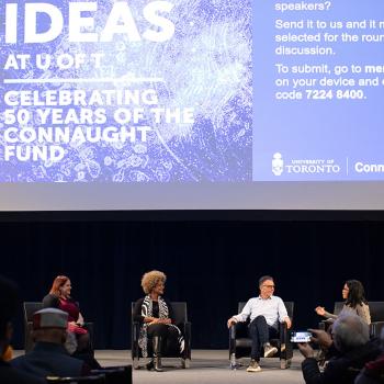 Four people sitting on stage speaking with one another, projection above them reads: Ideas at U of T. Celebrating 50 years of the Connaught Fund. 