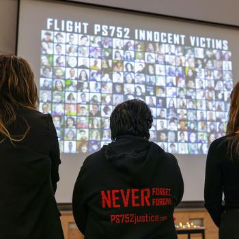 Three people stand with backs to the camera looking at a projection of faces on a screen. On the back of one coat is written: Never forget. Forgive. PS752justice.com