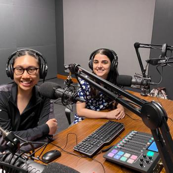Loridee De Villa and Harleen Kunda sit at a podcast booth, each wearing headsets with a midrophone in front of them. A computer keyboard, soundboard and monitor also sit on the wooden table in front of them.