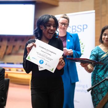 A graduate of the SEE UTM program in a black dress stands in front of officiants holding a certificate in front of her while smiling.