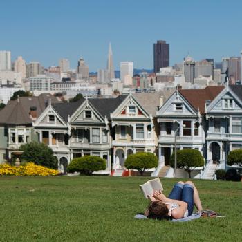 Woman laying in grass reading a book in a park, in background is row of colourful houses and farther back are taller buildings