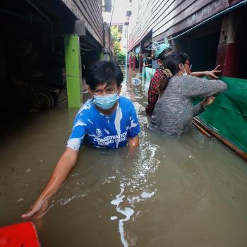 A man wearing a blue shirt and blue surgical face masks walks toward the camera in chest-high flood waters.rrow alley.