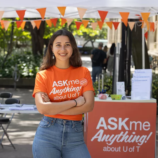 Ela Dai wearing an orange shirt that reads Ask Me Anything about U of T, standing in front of an outdoor tent and booth