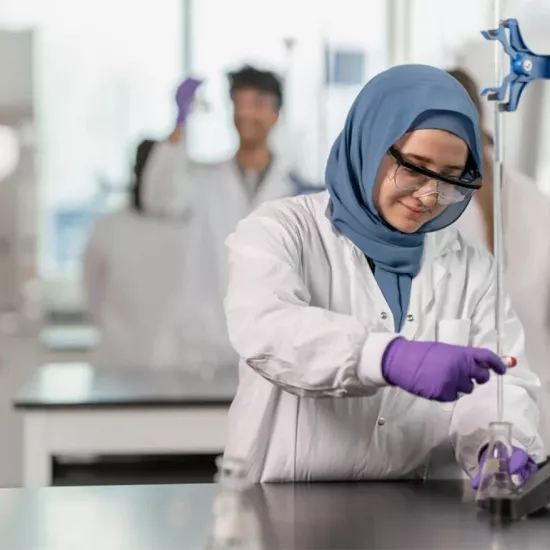 A woman in a lab coat wearing protective goggles measures a solution in a beaker