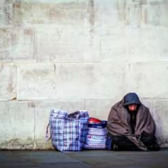 A homeless man sits on the ground. He is bundled in a blanket. Two large bags are beside him.
