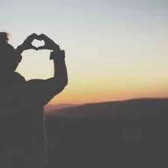 Woman holds hands in shape of a heart with sunset in the background