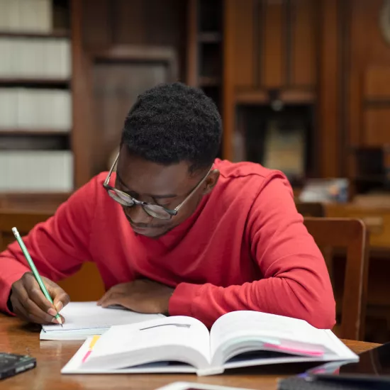 A student sitting at a desk in a library writing in a notebook.