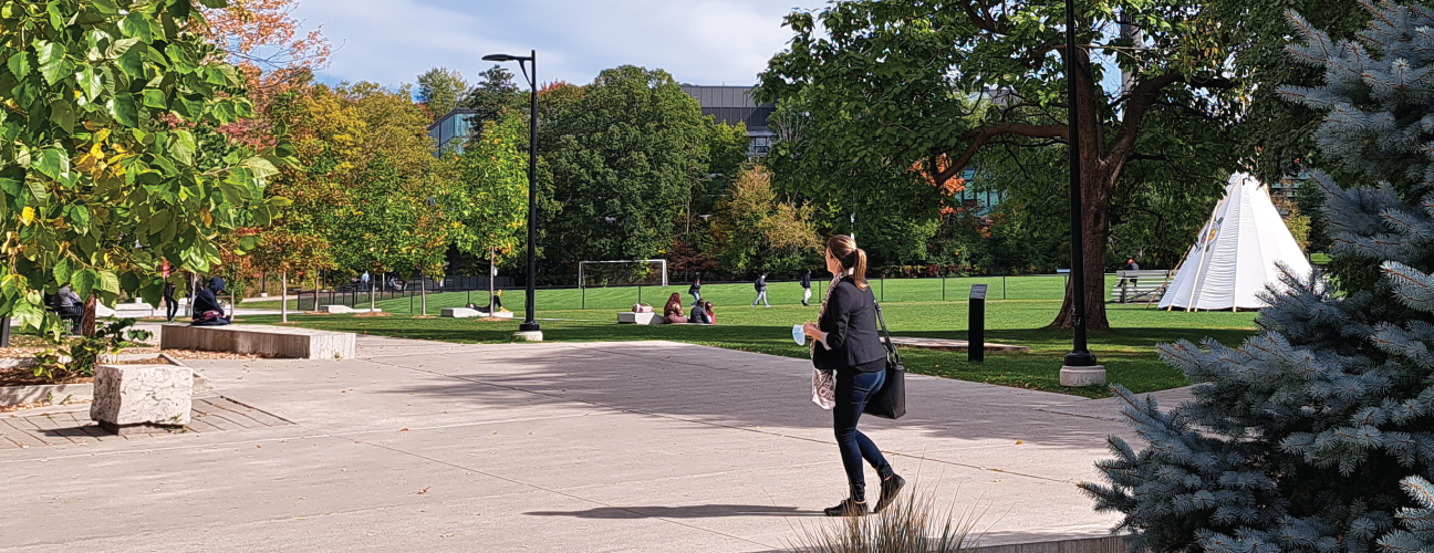 Photo of a student walking in front of Deerfield Hall with the Instructional building, campus green and UTM Tipi in the background