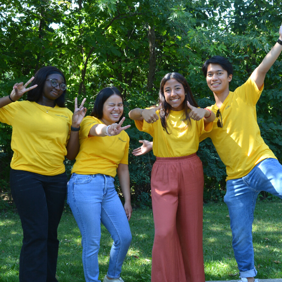 4 students in yellow shirts, outdoors, smiling and holding up peace signs.