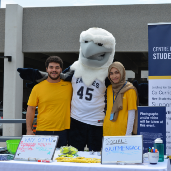 2 students in yellow shirts posing with the UTM Eagle mascot.