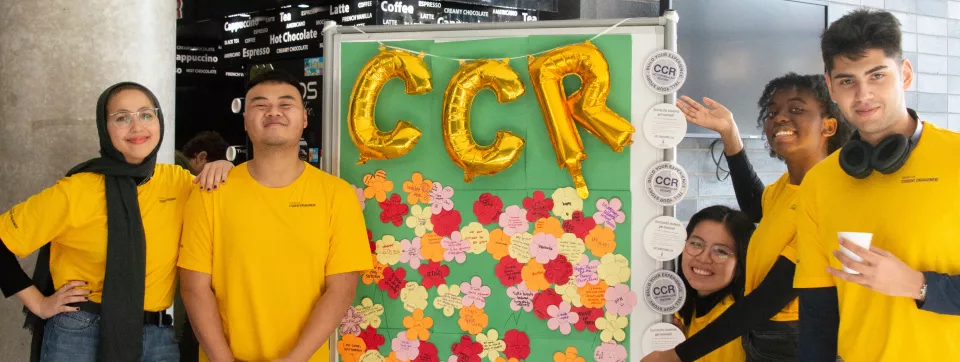 5 students smiling in front of a board that has balloons of the word "CCR."